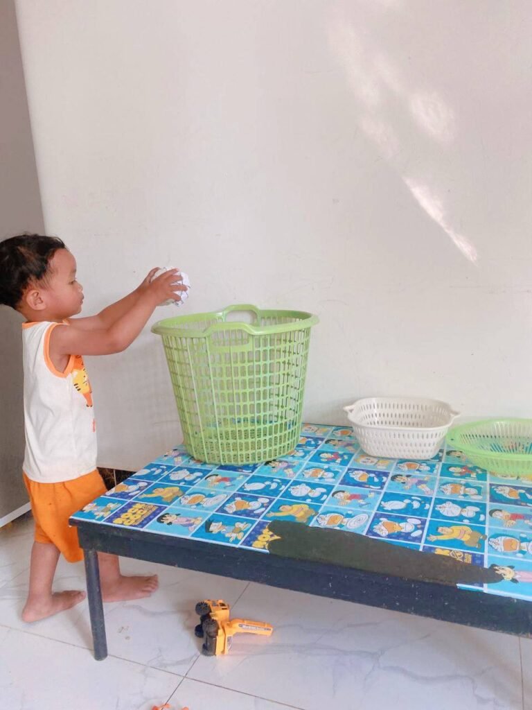 Toddler throwing a paper ball into a green laundry basket as part of a safe throwing activity.