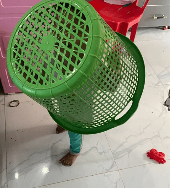 A toddler standing in a living room with a green plastic laundry basket stuck over their head.