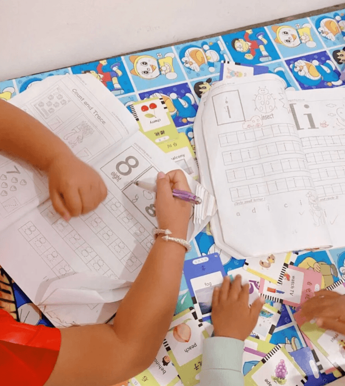A second grader tracing the number 8 and the word eight in a workbook with a purple pen, surrounded by English and Chinese vocabulary flashcards.