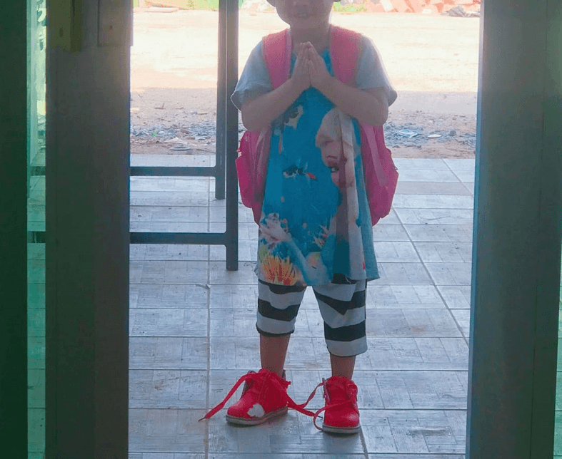 A young girl wearing a pink backpack practicing the traditional Cambodian Sampeah greeting at a doorway before leaving for school.