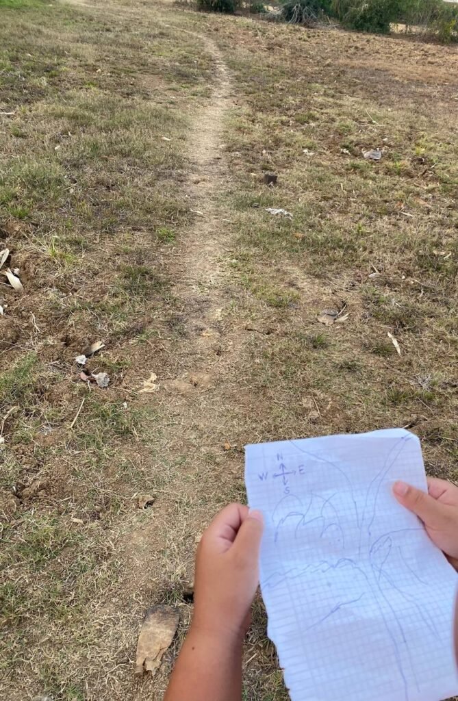 Child holding a hand-drawn map on grid paper with compass points on a dirt path in Cambodia.