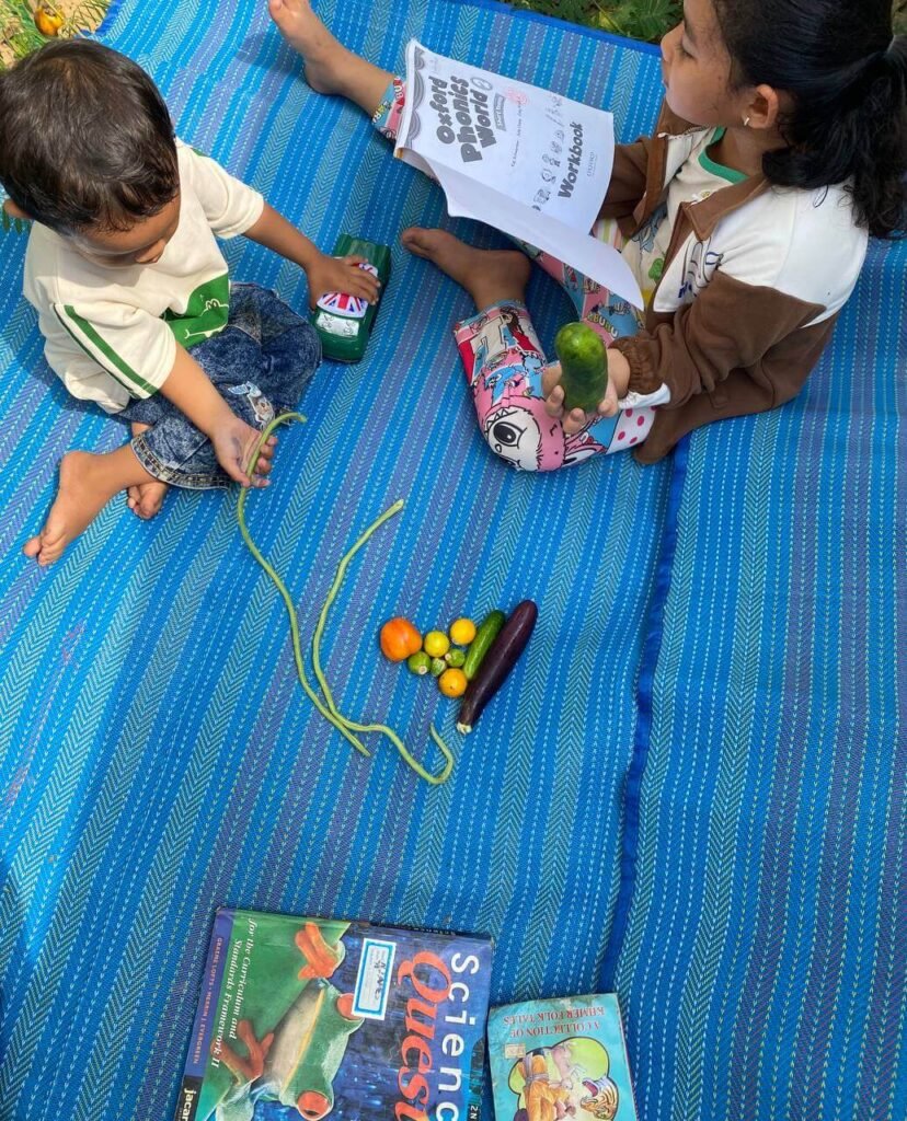 A young girl holding a fresh green cucumber in one hand while reading a phonics workbook to connect the physical object with the printed word.