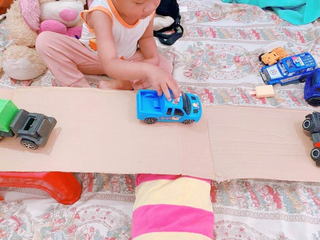 A toddler pushing a blue toy truck down a homemade DIY ramp made from folded cardboard.