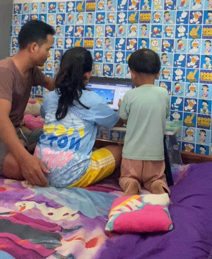 A family sitting together on a bed, playing an educational balloon typing game on a computer monitor to practice English.