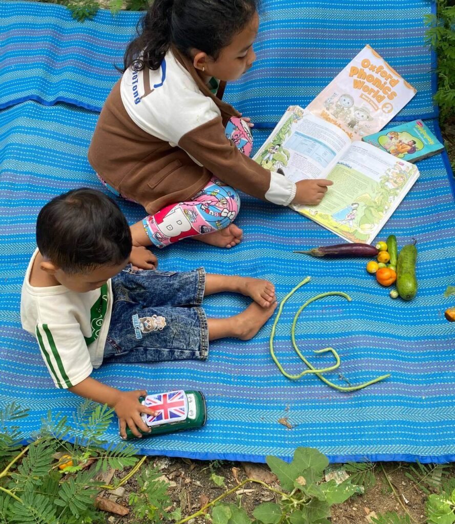 Two young children sitting on a blue woven mat in a garden, studying an Oxford Phonics workbook next to fresh vegetables.