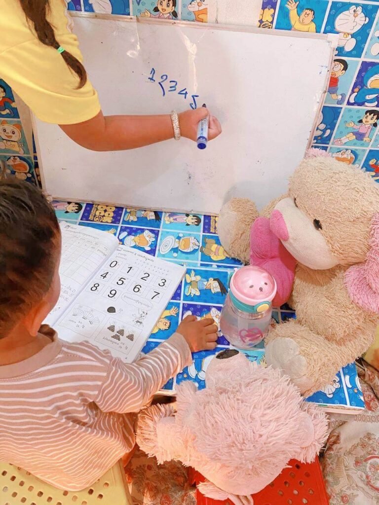 An older sister writing numbers on a whiteboard while her toddler brother looks at his matching exercise book.
