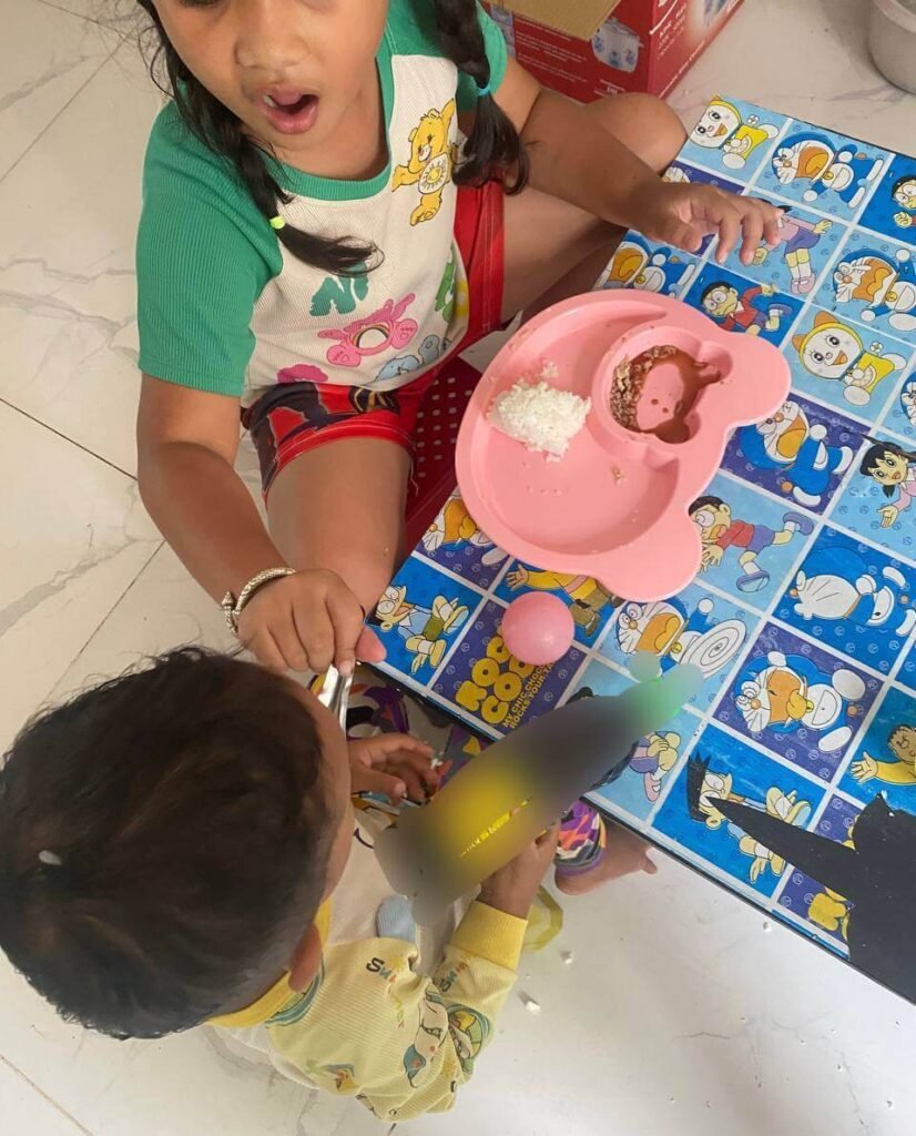 An 8-year-old older sister pretending to be a mom, successfully feeding her 2-year-old toddler brother rice from a pink plate.