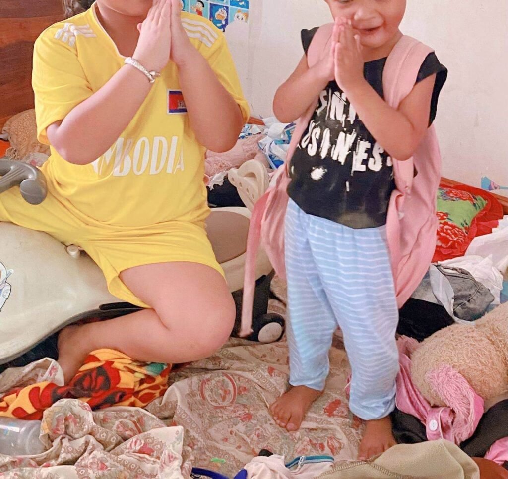 Older sister in a yellow Cambodia shirt teaching her toddler brother the traditional Cambodian Sampeah greeting while he wears a pink backpack.