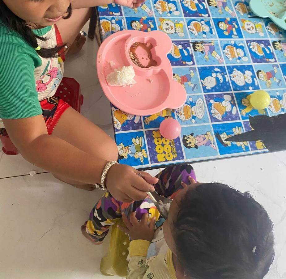 An older sister successfully feeding her toddler brother dinner, demonstrating peer-to-peer modeling for picky eaters.
