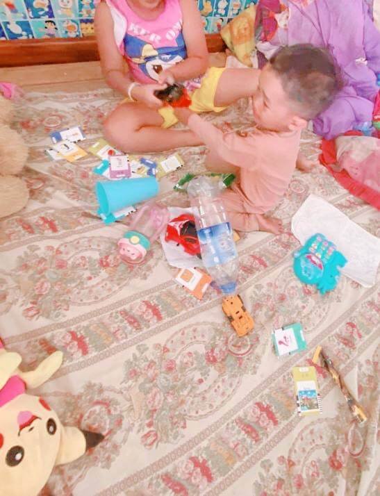 Toddler boy and older sister sitting on a floral floor mat pulling on a toy truck surrounded by flashcards.