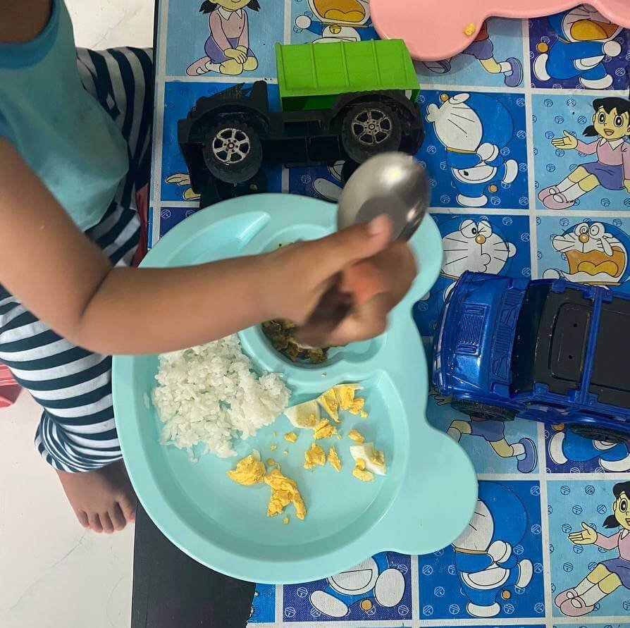 A toddler holding a spoon over a blue bear plate containing boiled duck eggs and rice, with a green toy truck nearby.