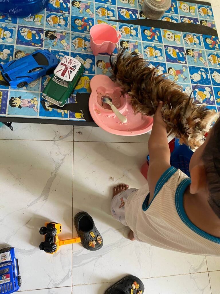 A toddler using a feather duster on a Doraemon table next to a pink bear plate and toy cars.