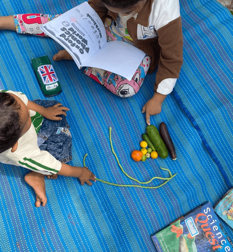 A toddler boy and his older sister pointing at fresh local vegetables like trub veng and long beans on a blue mat.