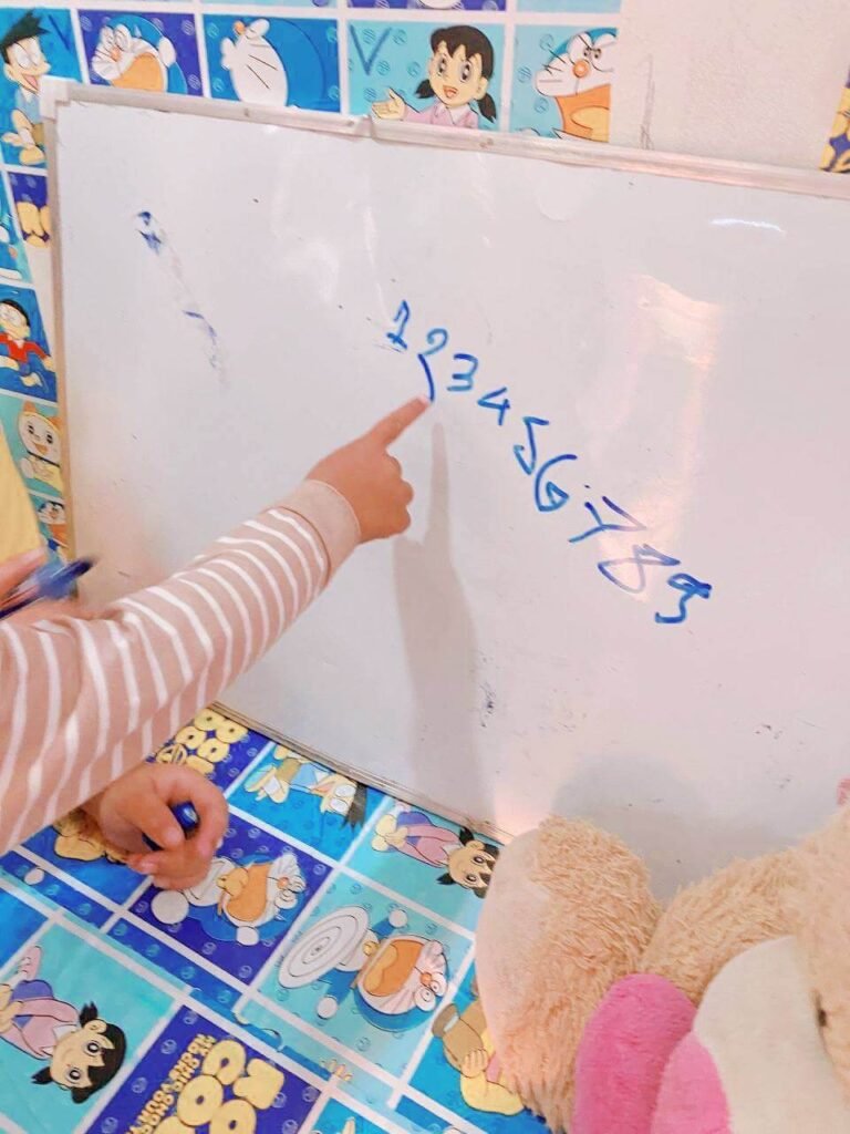 A toddler's hand pointing excitedly to the numbers 1 through 7 written in blue marker on a whiteboard.