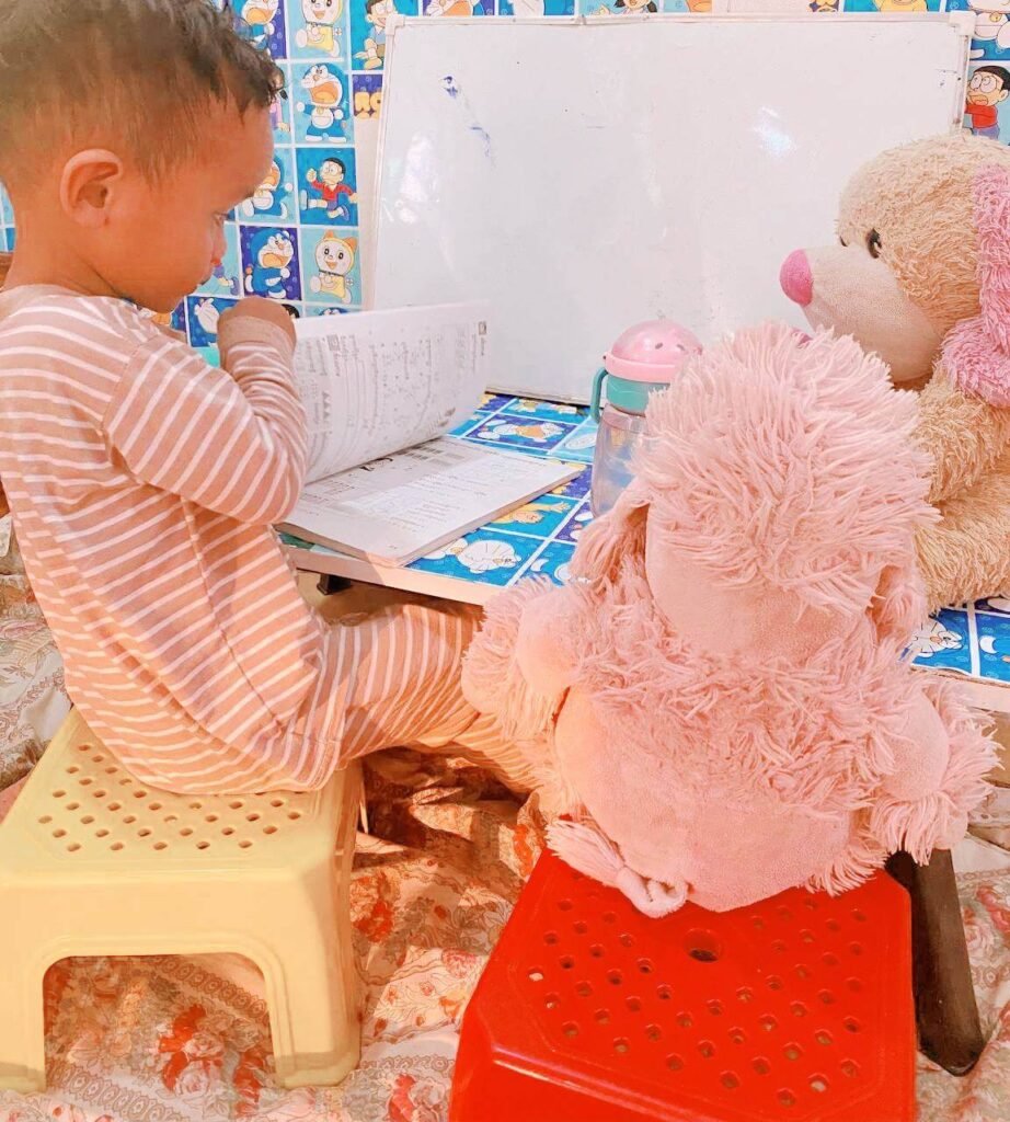 A toddler in striped pajamas looking at a math workbook at a small Doraemon table with stuffed animal classmates.