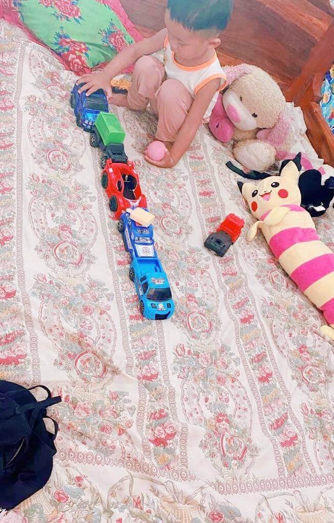 A toddler boy carefully lining up a row of colorful toy cars and trucks on a bed next to his plush toys.
