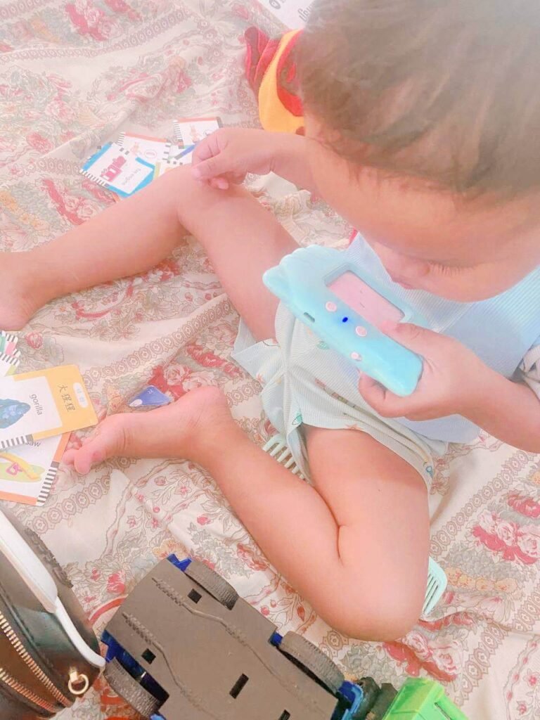 A toddler blowing into the slot of a blue reading machine toy to figure out how it works.