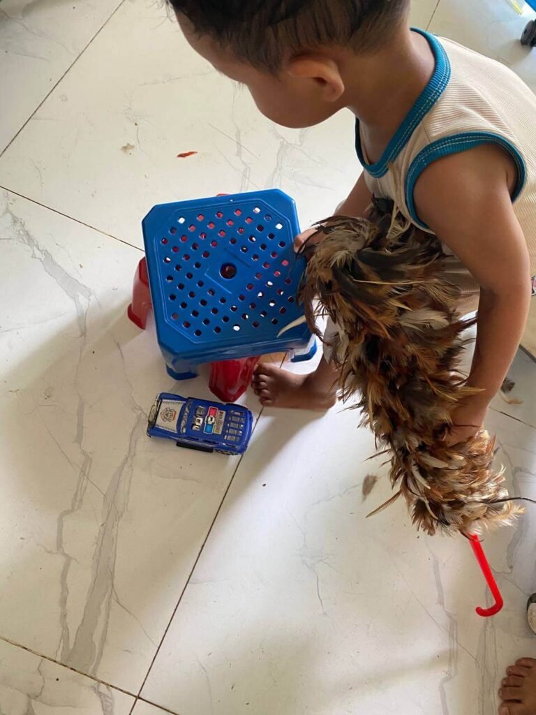 A toddler holding a feather duster while dragging a blue plastic stool across a white tile floor.