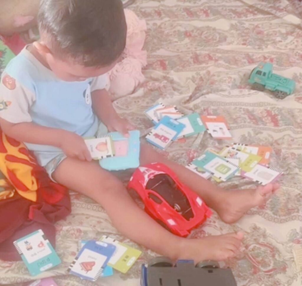 A toddler sitting on a floral blanket playing with scattered vocabulary flashcards and a blue reading machine.