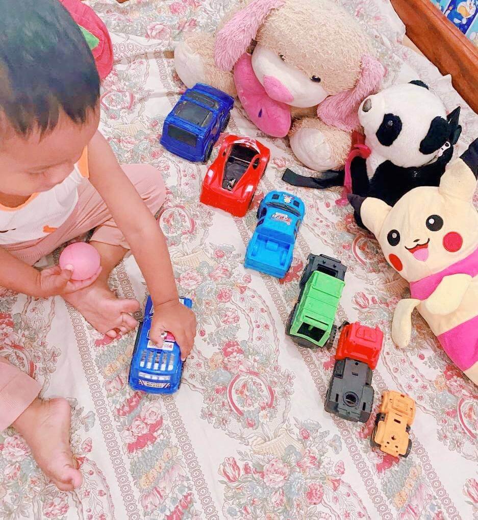Toddler boy sitting on a bed playing with a variety of toy trucks and cars, practicing his grip and hand-eye coordination.