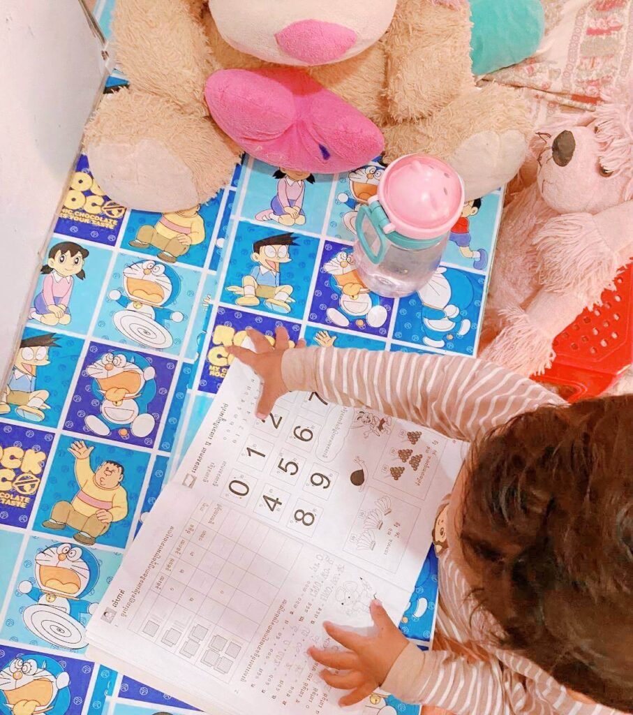 Top-down view of a toddler pointing at numbers in an open exercise book on a blue Doraemon table.