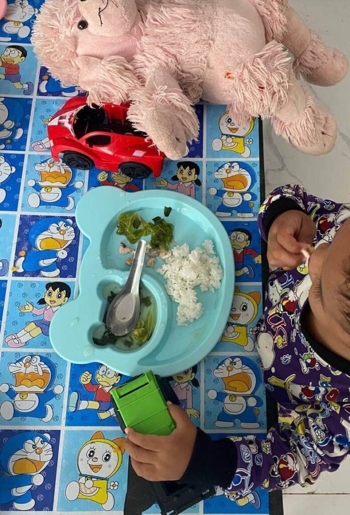 A toddler sitting at a table with a blue bear-shaped plate of rice and greens, surrounded by a pink fluffy toy and a red toy car.