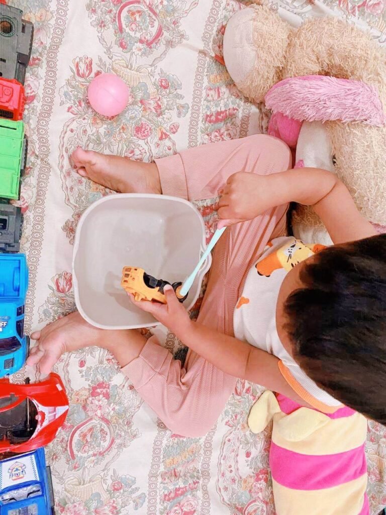 A toddler boy focused on scrubbing a yellow toy car with a toothbrush in a small grey basin of water.