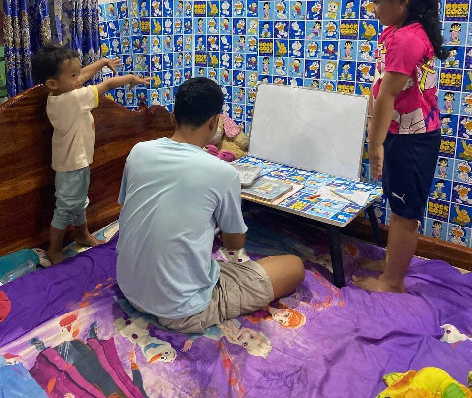 A father and two children sitting on a bed looking at a visual routine whiteboard for kids.