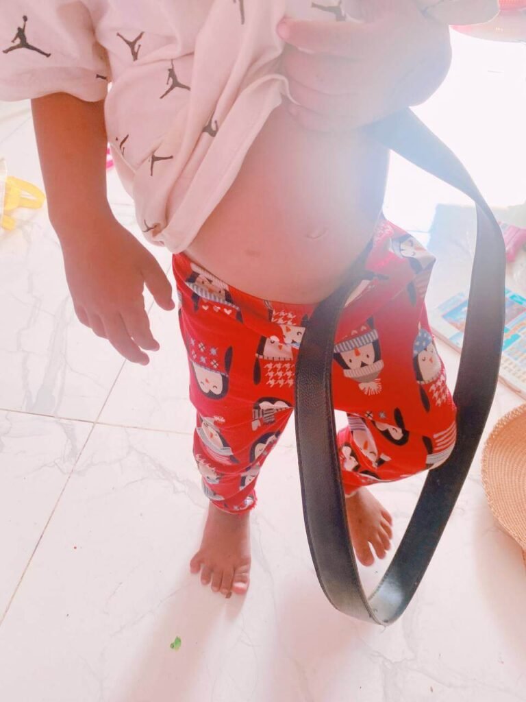 A toddler looking down at an oversized adult belt draped around them, practicing habits learned through observation.