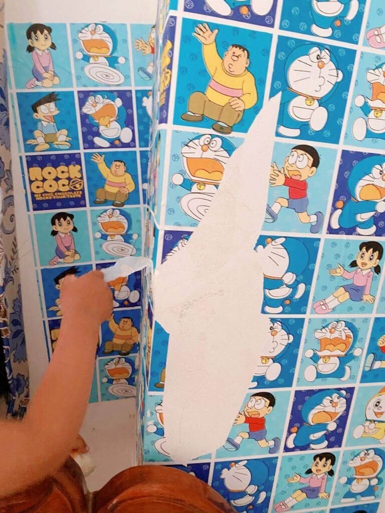 A hand peeling away a section of blue wallpaper, revealing the bare white wall underneath.