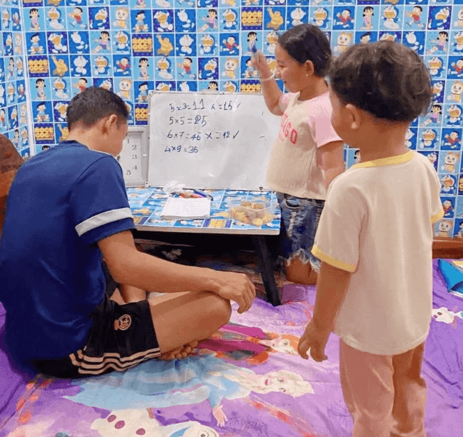 A father and daughter practicing math on a small whiteboard while a toddler watches, surrounded by blue Doraemon wallpaper.