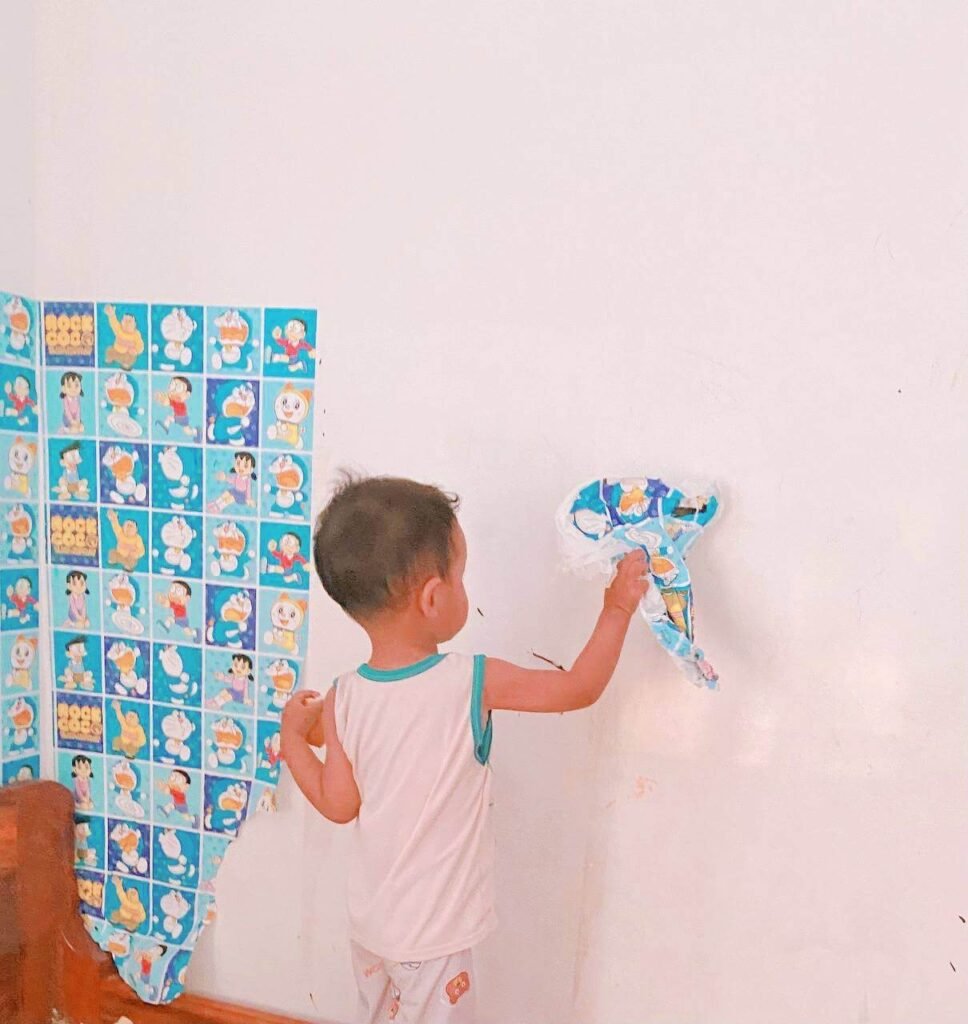 A toddler standing next to a partially stripped wall, holding a crumpled piece of the torn wallpaper.