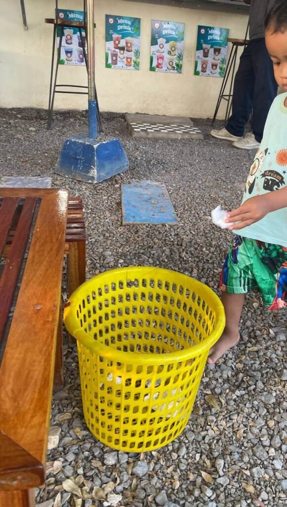 A barefoot toddler walking across an uncomfortable gravel surface to throw a tissue into a yellow trash bin.
