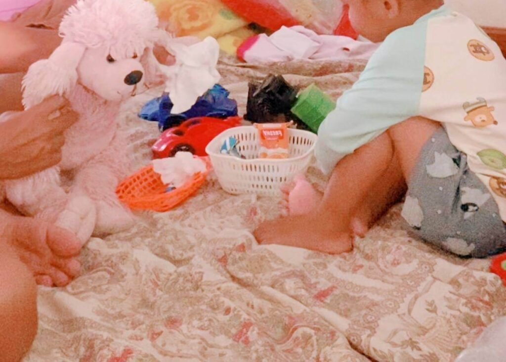 An adult holding a pink poodle stuffed animal, demonstrating how to throw away a tissue for a toddler playing on a bed.