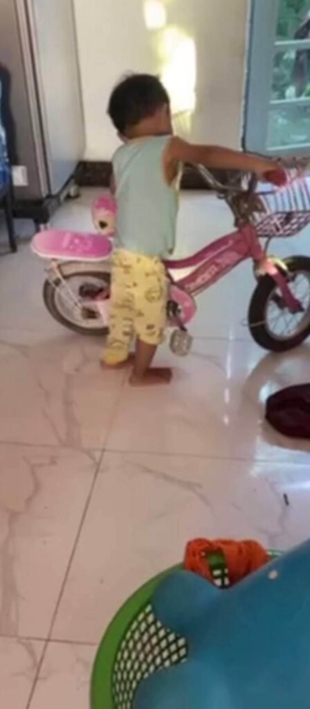 A toddler standing indoors next to an older sibling's pink bicycle.