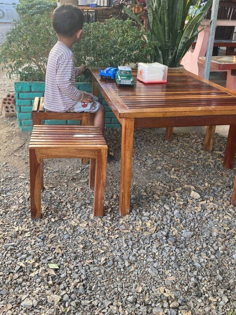 A toddler sitting at a wooden table at an outdoor café, playing with toy cars next to a gravel path.
