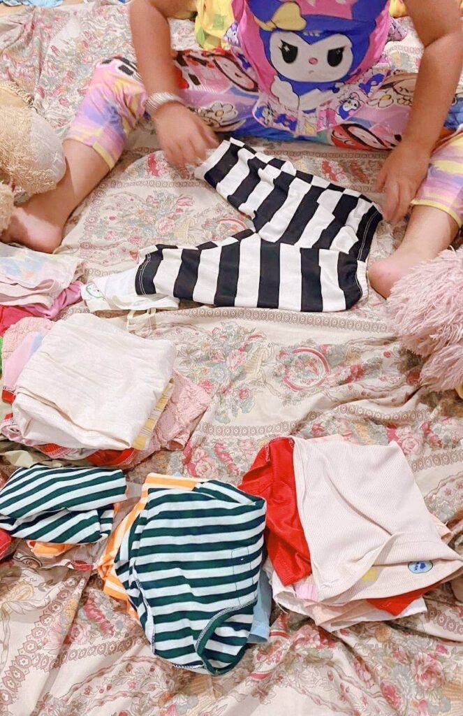 A young girl wearing a Kuromi shirt sitting on floral bedsheets, neatly folding black and white striped pants.