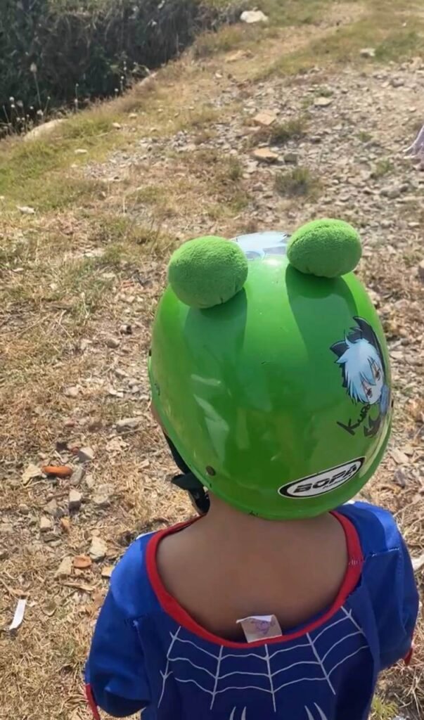 A toddler standing outdoors wearing a bright green bicycle helmet with plush pompom ears.