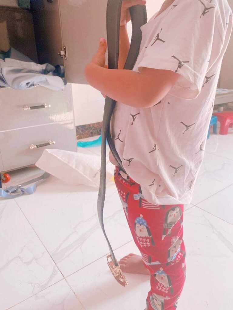 A toddler in pajamas holding a large adult leather belt, illustrating how children observe parent routines.