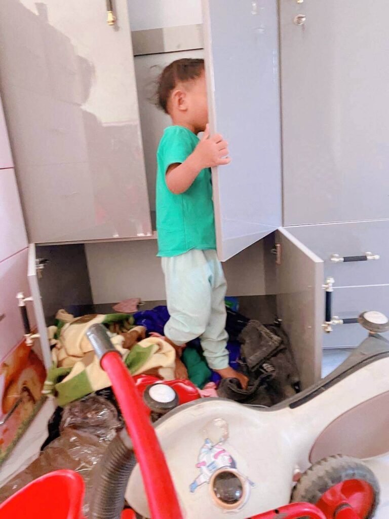 A two-year-old boy standing inside a new, empty grey wardrobe, looking into the open cabinet doors.