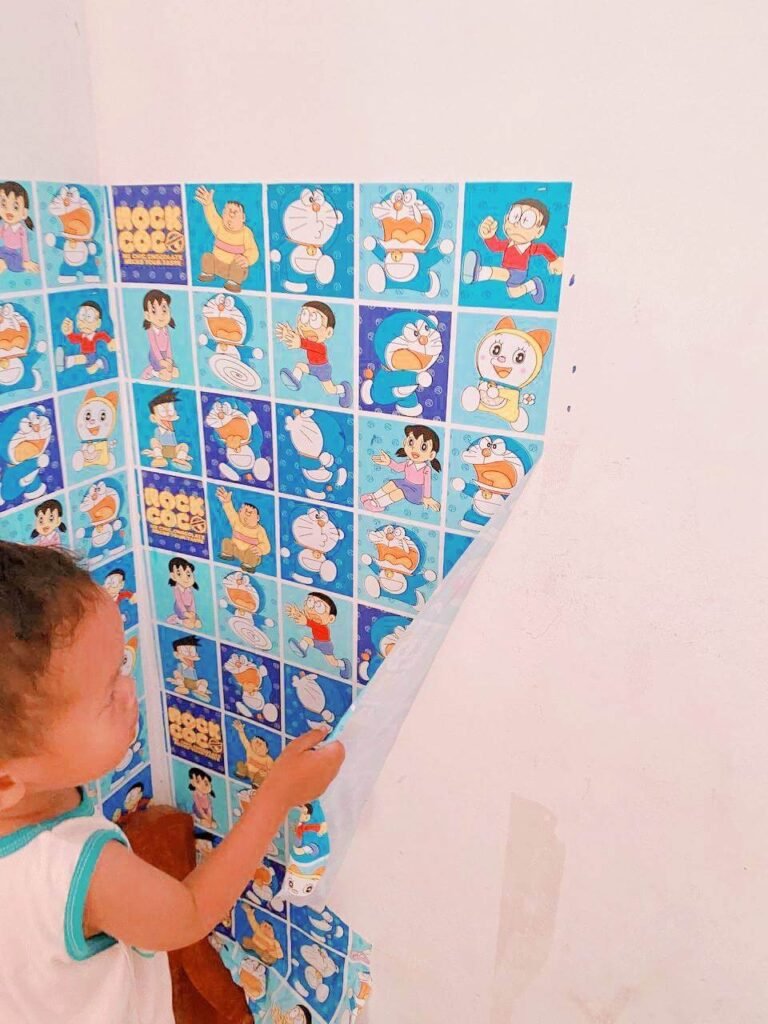 A young toddler actively peeling a large sheet of Doraemon wallpaper away from the bedroom wall.