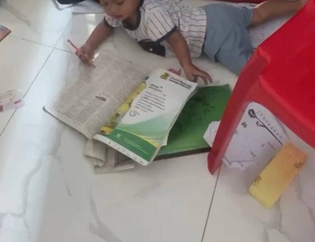 A toddler lying on a tile floor scribbling with a pencil inside an open workbook.