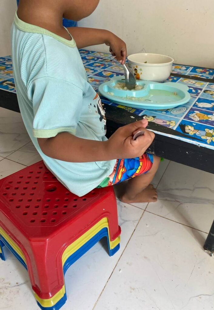 A toddler eating breakfast while sitting on a tall stack of red, yellow, and blue plastic stools.