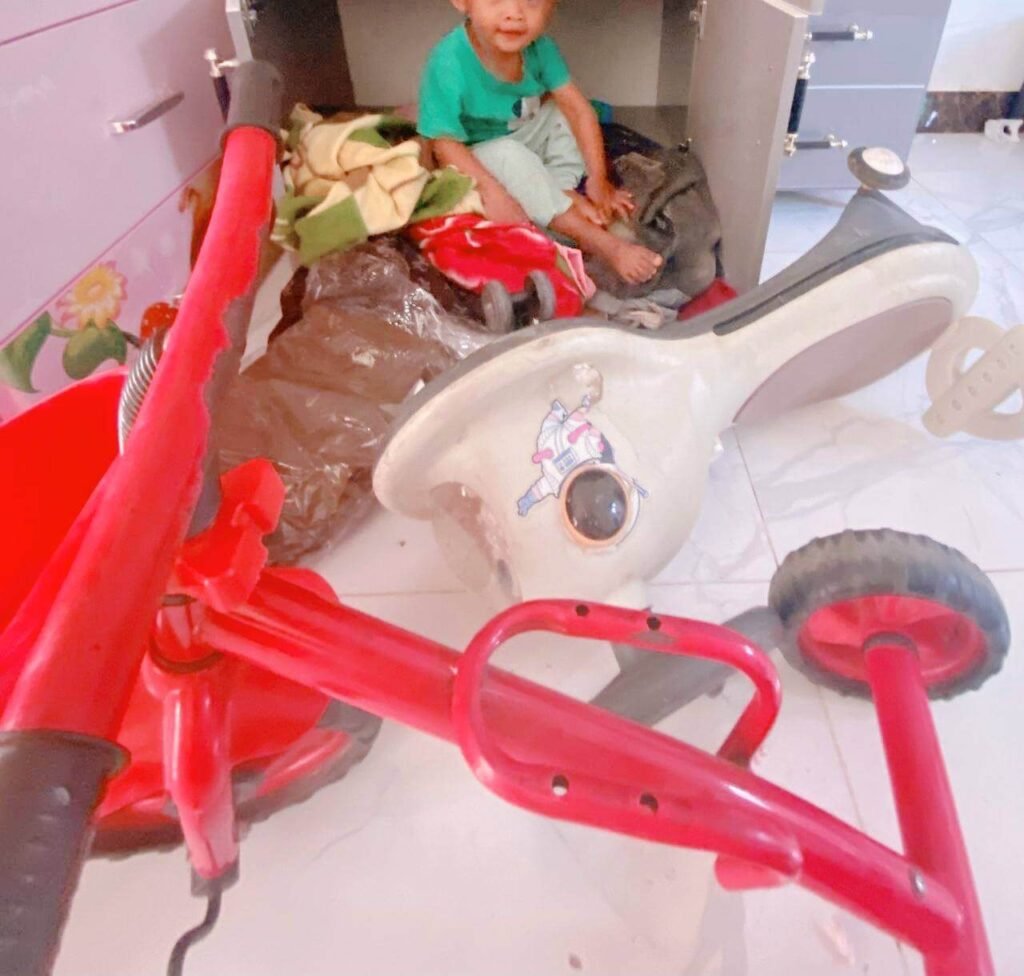 A toddler boy protesting by sitting on blankets inside the bottom of a grey wardrobe, sitting behind a tipped-over red tricycle.
