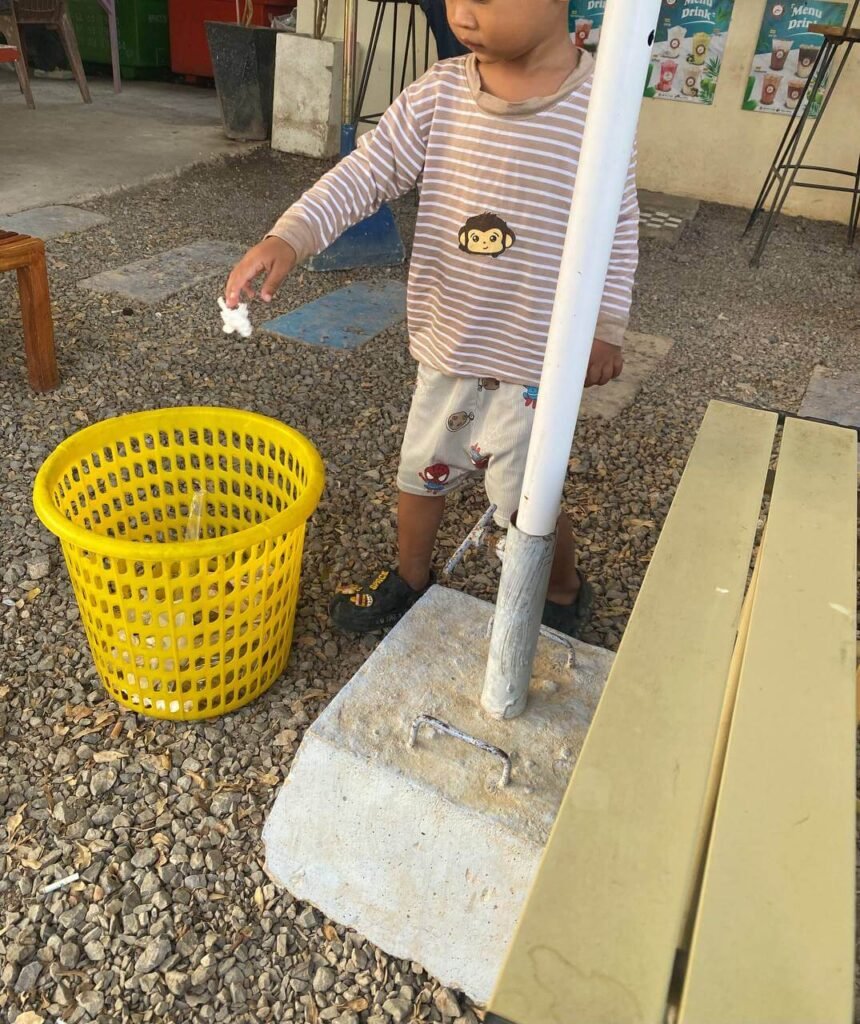 A toddler wearing shoes standing next to a gravel area, successfully dropping a tissue into a yellow plastic trash bin.