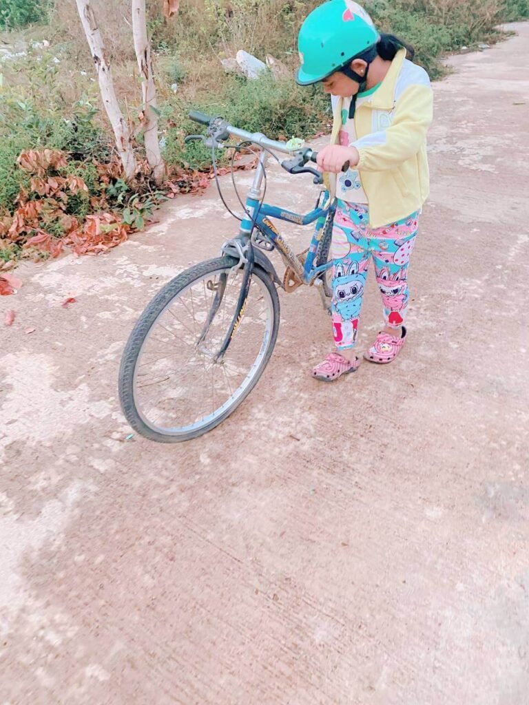 An 8-year-old girl wearing a green safety helmet and yellow jacket carefully inspecting a large adult bicycle on a concrete path.