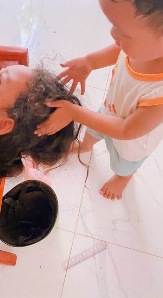 A 2-year-old toddler boy standing and placing his hands in the messy hair of his older sister, who is sitting on a wooden bench.