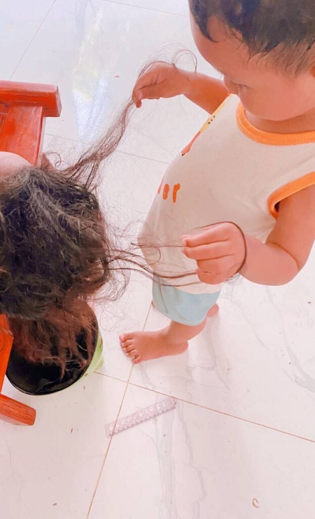A toddler boy carefully holding up a strand of his sister's hair during a pretend play hair salon session at home.
