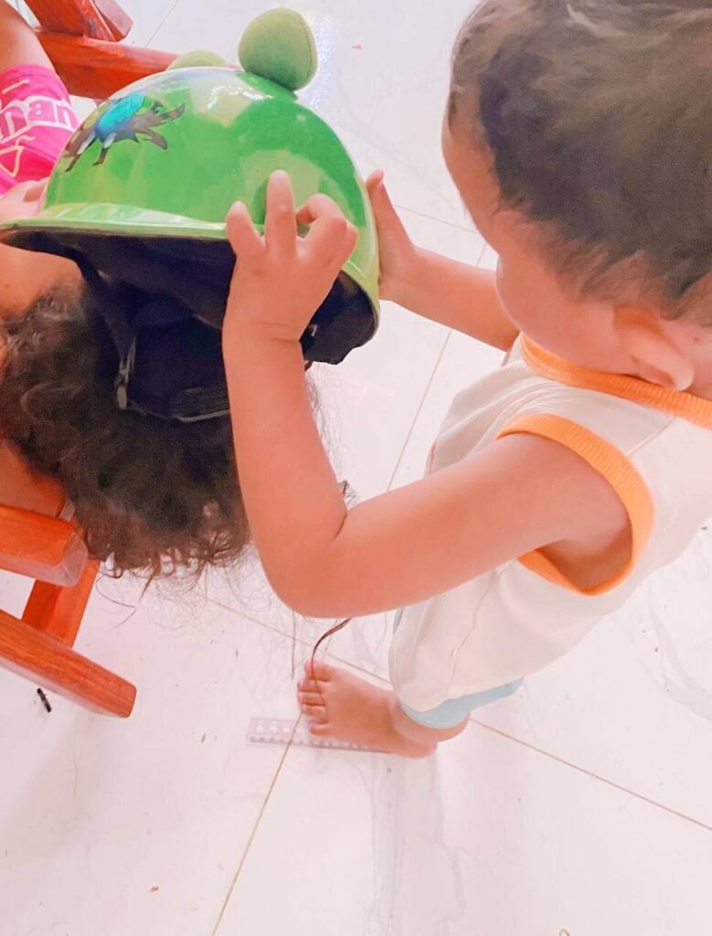 A toddler boy placing a bright green frog helmet with eyes onto his older sister's head after pretending to wash her hair.